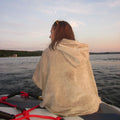 A woman sitting on a boat, wrapped in a light beige hooded towel poncho, looking out over a lake at sunset.