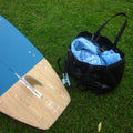 A blue and wood paddle board is placed on green grass next to a black bag containing a light blue TAIGA BOARD Surfing towel.