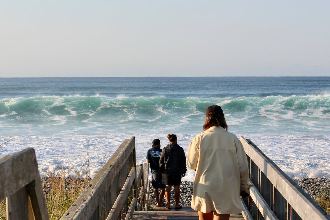 Three people walking down a wooden staircase towards the ocean with waves crashing below.