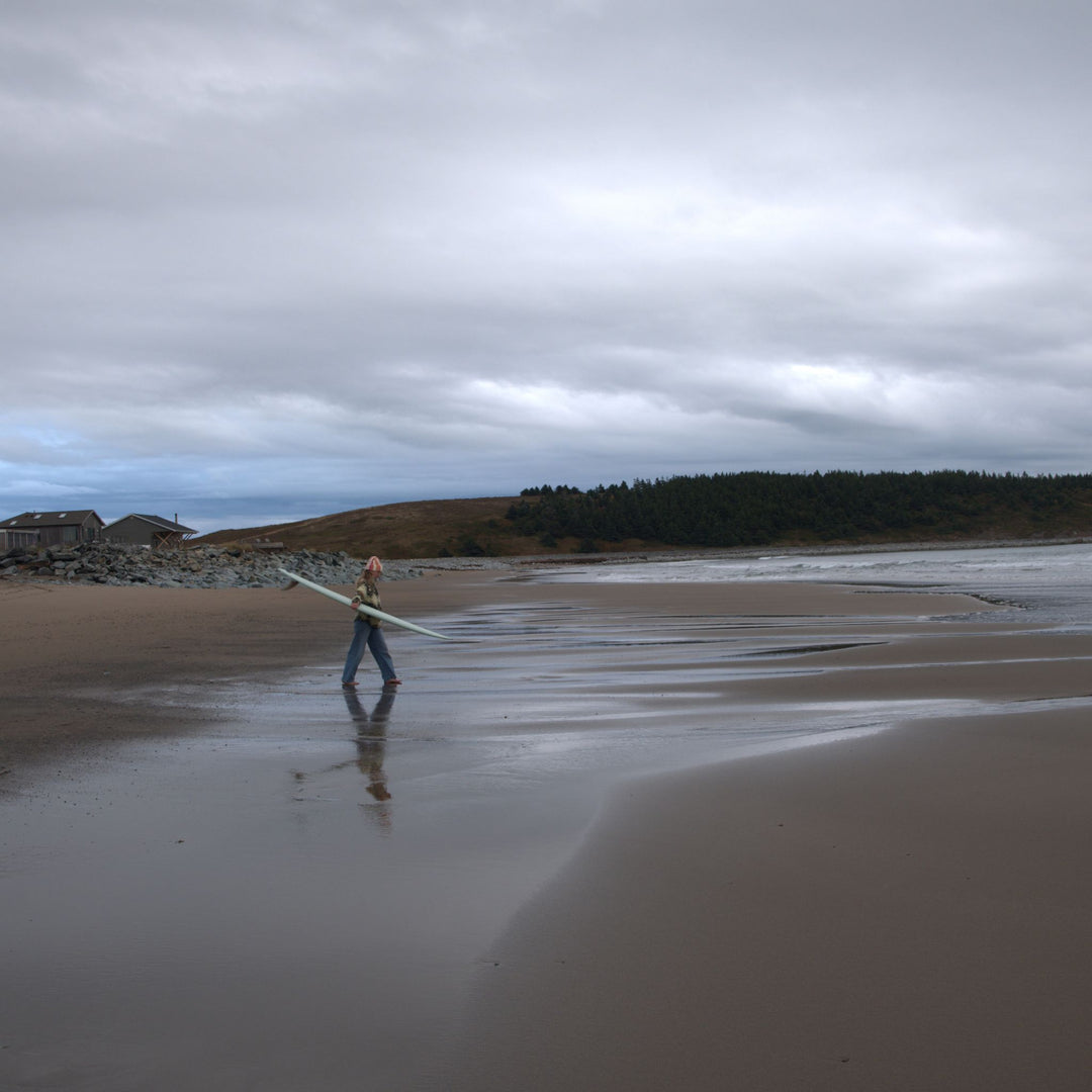 Person walking on a beach with a longboard, under a cloudy sky.