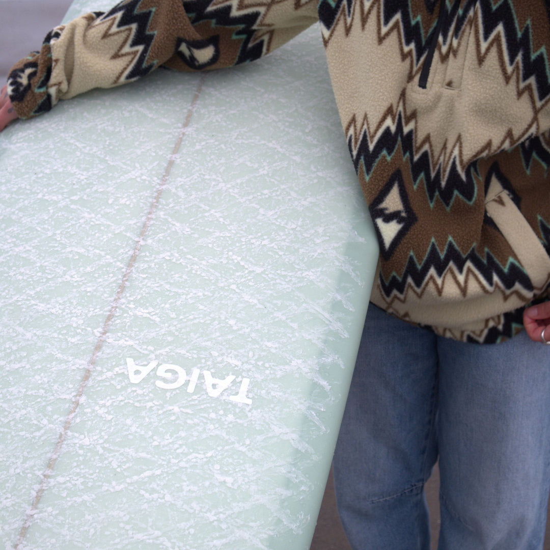 Person wearing a patterned sweater and blue jeans standing next to a Taiga surfboard