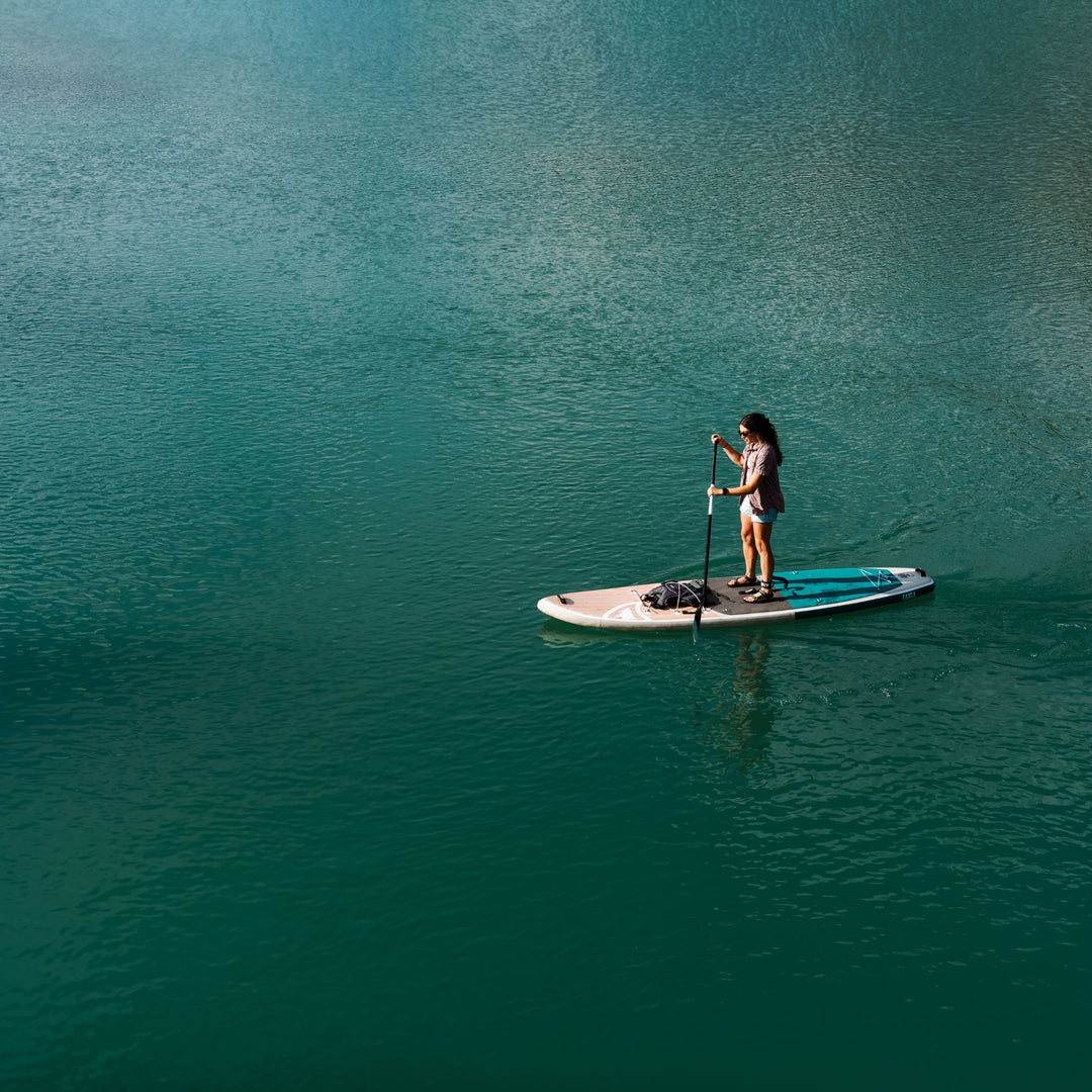 A person stands on a paddleboard on calm turquoise water, holding a paddle. The board is white with blue and black details, and the water fills the frame.