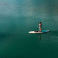 A person stands on a paddleboard on calm turquoise water, holding a paddle. The board is white with blue and black details, and the water fills the frame.