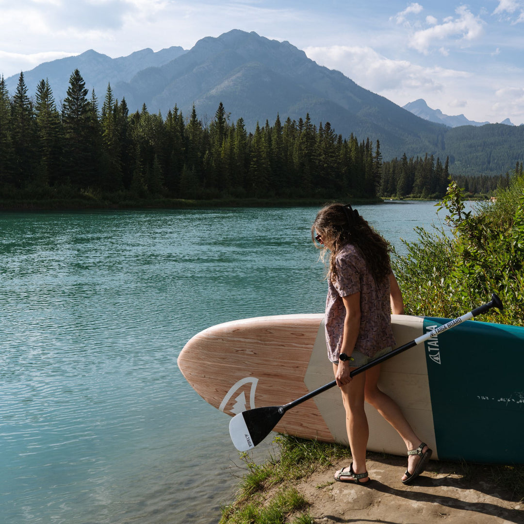 A woman stands on a lakeshore holding a paddle and a paddleboard, with a scenic mountain and forest background.