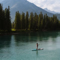 A person stands on a paddleboard on a calm turquoise lake, with a dense evergreen forest and mountains in the background.