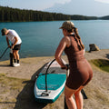 Two people with a paddleboard on a lakeside with trees and mountains in the background.