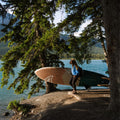 Person holding a paddle board by a lake with trees and mountains in the background