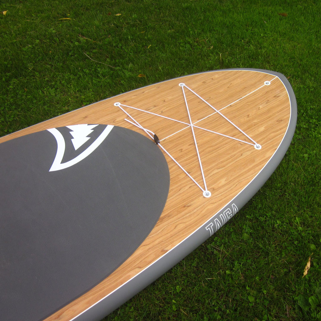 A stand-up paddle board photographed outdoors on grass, showing a natural wood deck, dark gray pad, white bungee cords, and a gray edge.