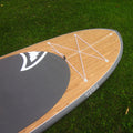 A stand-up paddle board photographed outdoors on grass, showing a natural wood deck, dark gray pad, white bungee cords, and a gray edge.