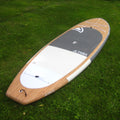 A Taiga hard stand-up paddle board lying on green grass, showing the natural wood deck, white and gray pad sections, and the Taiga brand name.