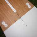 The underside of a wooden outdoor table on a grassy lawn, showing a light wood grain top, white plastic mounting brackets, and handwritten dimensions reading 'AWEN 10' x 33" x 4.7" 191L'.