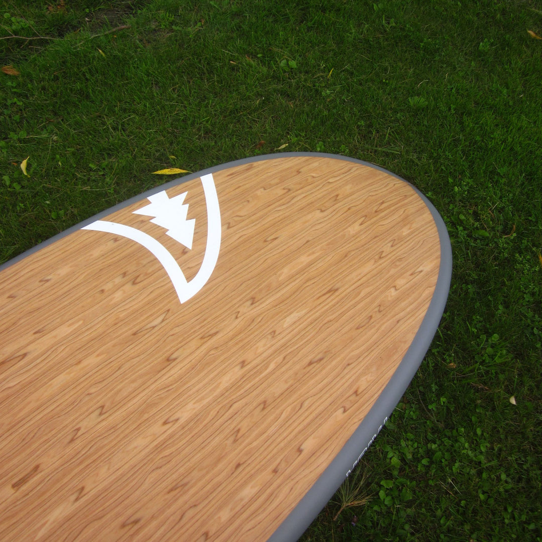 A close-up, angled view of a paddleboard deck featuring a light brown wood grain finish and a white logo, resting on green grass.