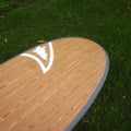A close-up, angled view of a paddleboard deck featuring a light brown wood grain finish and a white logo, resting on green grass.
