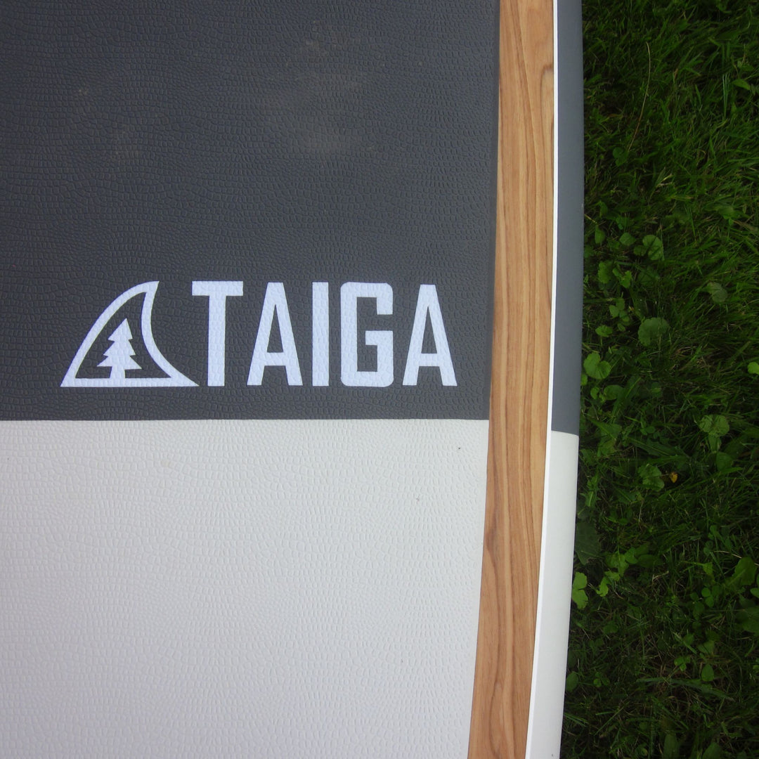 Close-up of a Taiga paddle or board section showing a dark gray textured section with the white TAIGA logo, a light wood edge, and part of a white lower section, photographed outdoors next to green grass.