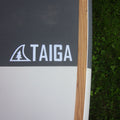 Close-up of a Taiga paddle or board section showing a dark gray textured section with the white TAIGA logo, a light wood edge, and part of a white lower section, photographed outdoors next to green grass.