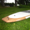 A wooden paddle board lying on green grass in an outdoor park setting, with trees and water visible in the background. The board has a natural wood nose, a white deck, and a dark gray tail section with visible branding.
