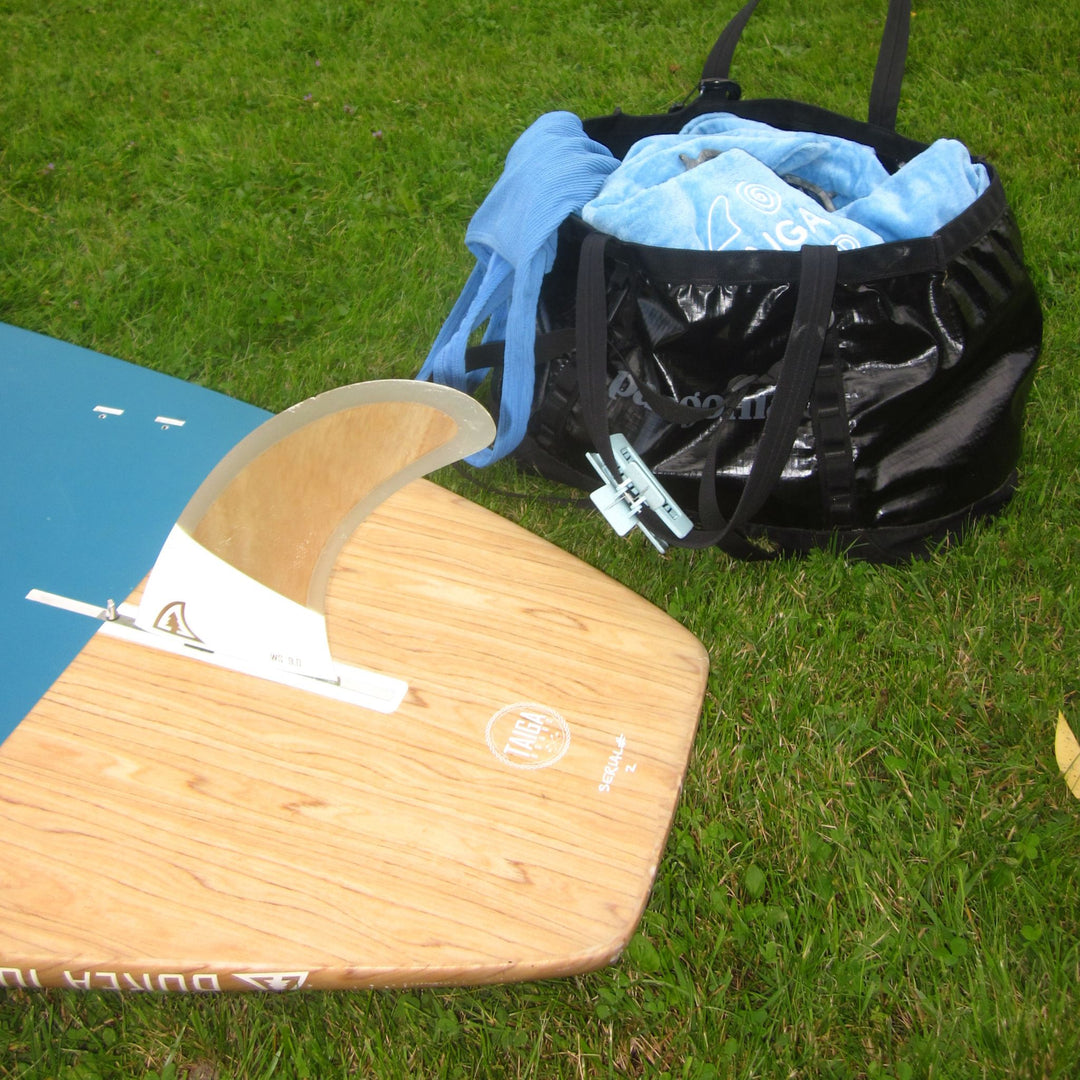 A wooden surfboard with a blue tail and white fin is laid on grass next to a black duffel bag containing a light blue chaging robe.