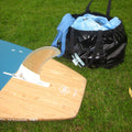 A wooden surfboard with a blue tail and white fin is laid on grass next to a black duffel bag containing a light blue chaging robe.