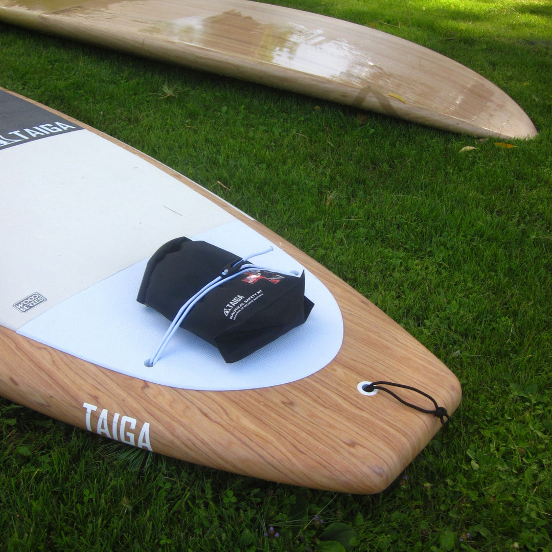 A wooden surfboard with a white deck pad and a black ankle leash lies on green grass, with another surfboard blurred in the background.