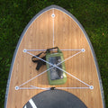 An overhead view of a paddleboard with a light wood-grain deck, gray rails, white bungee cords, and a green waterproof dry bag mounted on the deck, placed on grass.