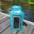 A turquoise blue Taiga waterproof dry bag with a clear front window, standing on a wooden dock with water and greenery in the background.