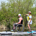 Two people on an Adaptive Inflatable Paddle Board with stabilizers in calm water.