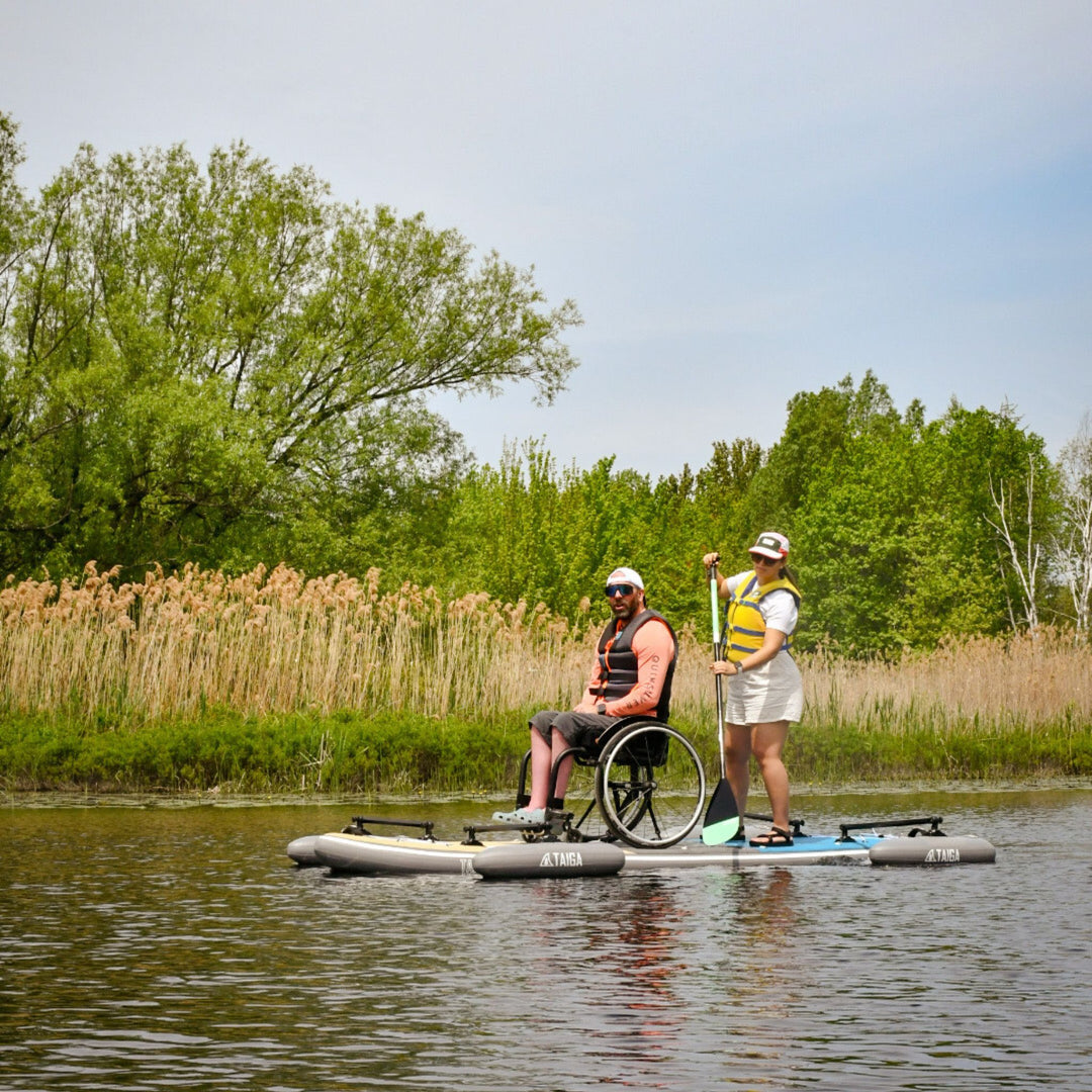 Two people on a paddleboard with one in a wheelchair, surrounded by trees and reeds.