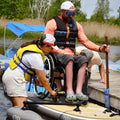 Person in a wheelchair being assisted by another person on a dock with water and trees in the background