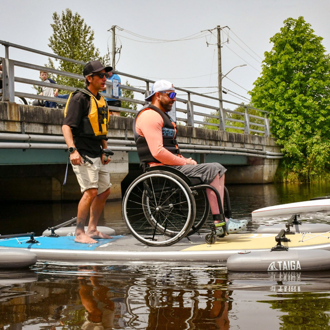Two people on a paddleboard with one in a wheelchair, on a calm body of water.