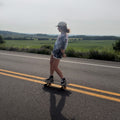 A person stands on a skateboard on a paved road with a green field and trees in the background, wearing a gray patterned t-shirt, white shorts, and black sneakers.