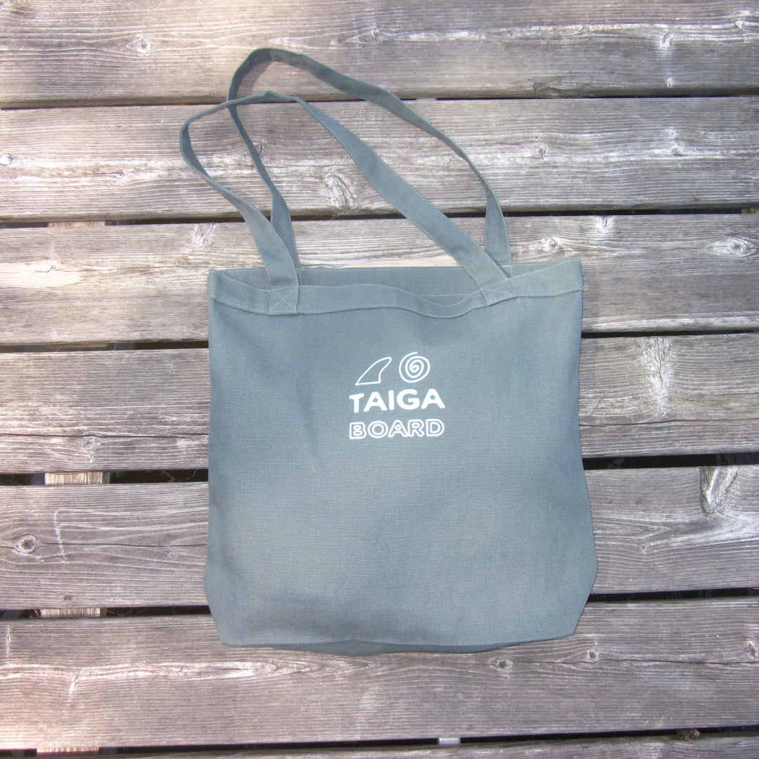 A teal canvas tote bag with white printed text reading 'TAIGA BOARD' and a small logo, placed on a weathered gray wooden plank surface.