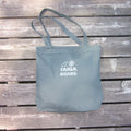 A teal canvas tote bag with white printed text reading 'TAIGA BOARD' and a small logo, placed on a weathered gray wooden plank surface.