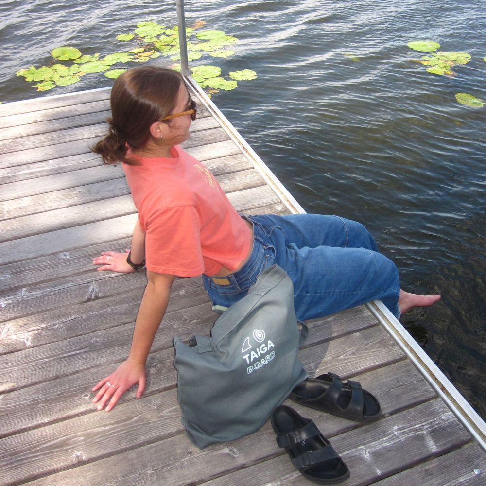 A person sitting on a wooden dock by the water, wearing a coral t-shirt, blue jeans, and black sandals, with a teal TAGA bag beside them. Lily pads are visible in the water.