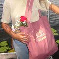 A person outdoors by the water carries a dusty pink canvas tote bag with the words 'TAIGA BOARD' printed in yellow. The person is wearing a light beige t-shirt and blue jeans, holding a small bouquet of red and white flowers.
