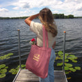 A woman stands on a wooden dock by a lake, seen from behind, carrying a dusty pink canvas tote bag with gold 'TAIGA BOARD' branding. The tote bag has a small red flower tucked into one of its straps.