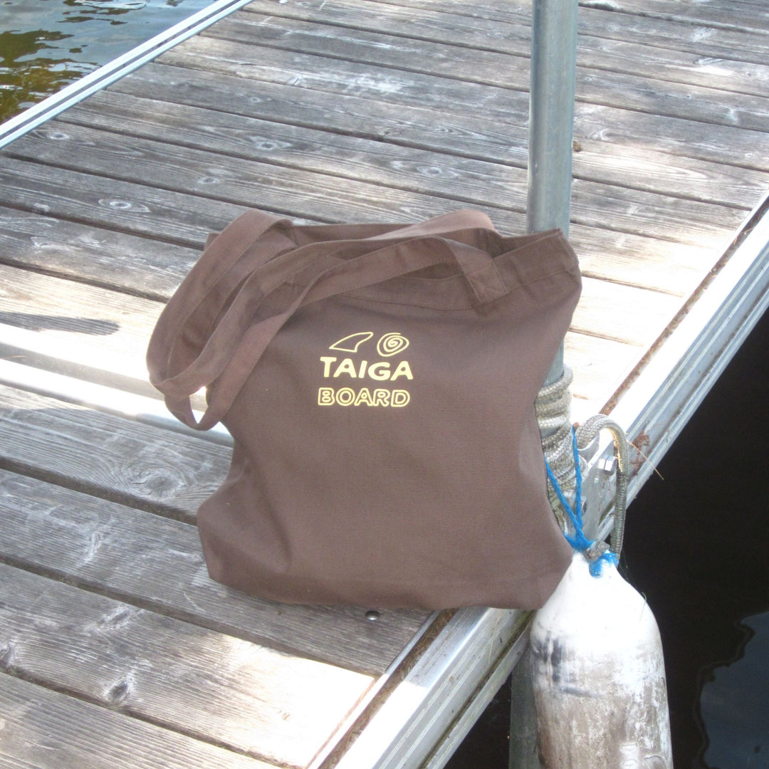 A brown canvas tote bag with 'TAIGA BOARD' printed in gold on the front, resting on a weathered wooden dock next to water.
