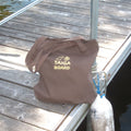 A brown canvas tote bag with 'TAIGA BOARD' printed in gold on the front, resting on a weathered wooden dock next to water.