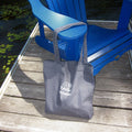A marine tote bag with white printed text and logo sits on a weathered wooden dock next to a bright blue Adirondack chair by the water.
