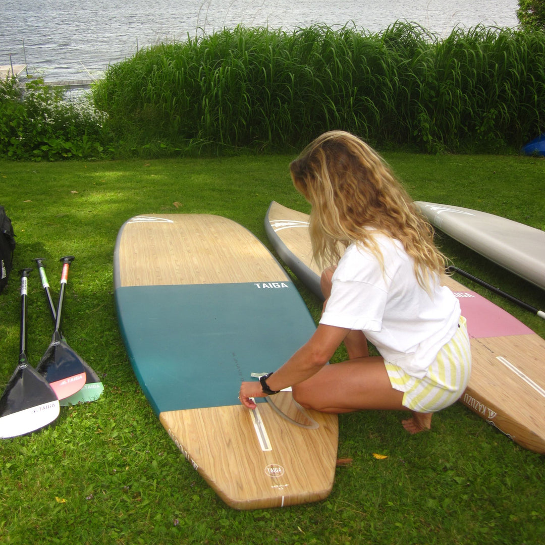 A woman installs the fin of a wooden and teal stand-up paddle board, with other paddle boards and paddle blades nearby and water and reeds in the background.