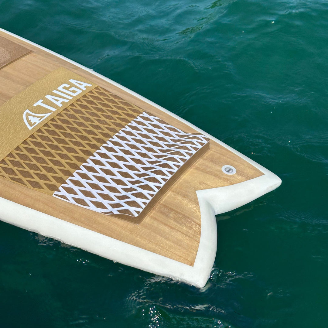 Close-up of a Taiga wakesurf floating on turquoise water, showing the wood-grain deck, traction pad, and white edge.