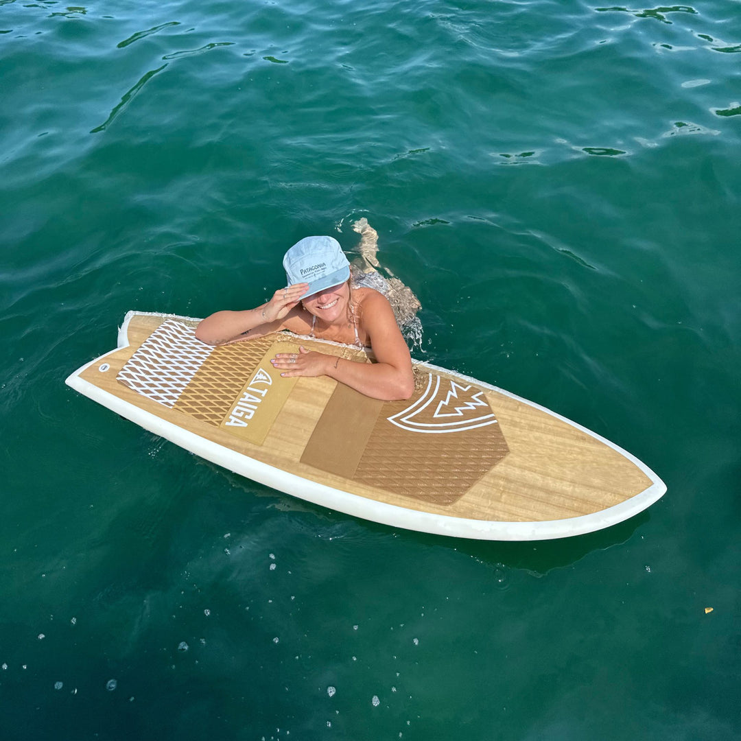 A woman wearing a light blue cap lies on a wooden wakesurf in the ocean, with the brand name 'TAIGA' visible on the board.