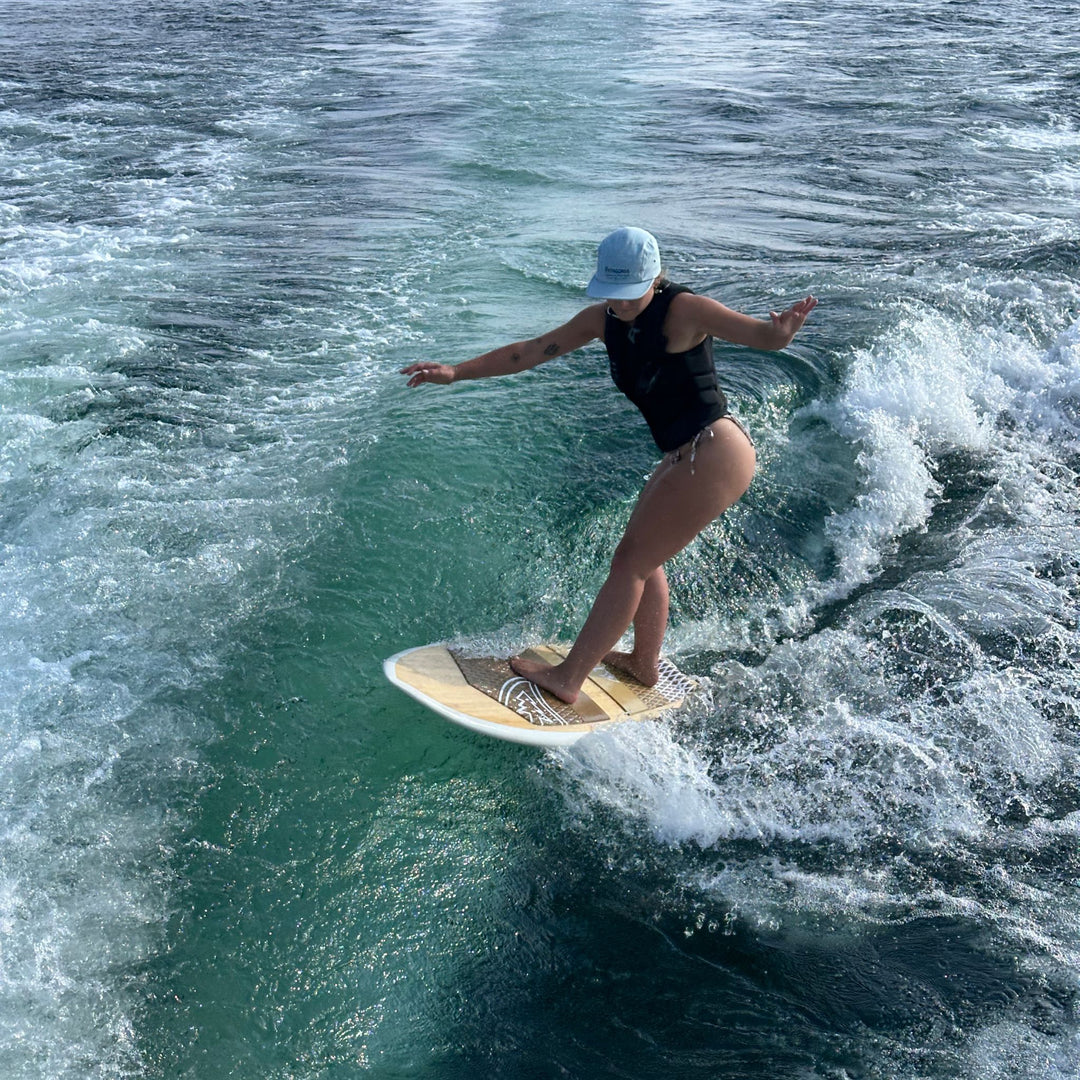 A woman in a black swimsuit and light blue cap stands on wakesurf riding the wake of a boat in blue-green water.