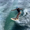 A woman in a black swimsuit and light blue cap stands on wakesurf riding the wake of a boat in blue-green water.